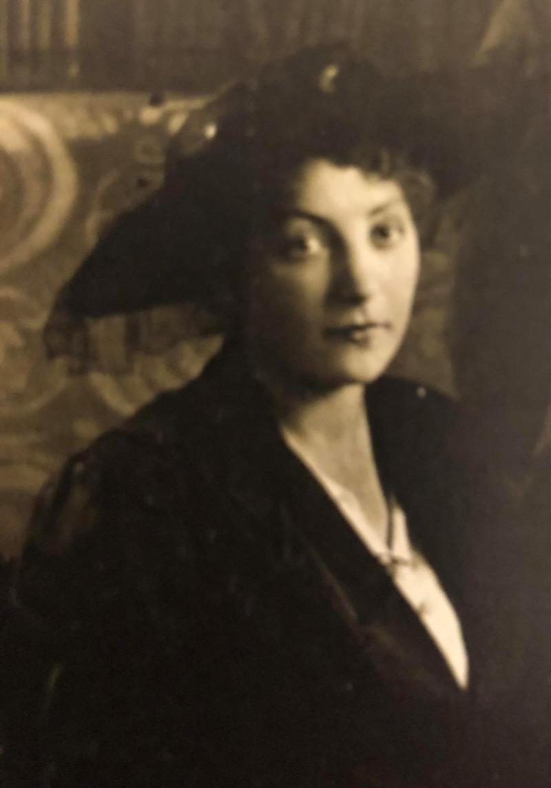 Die monochrome Fotografie in Sepiatönen zeigt Anna le Coutre. Ein junge Frau mit Hut und welligem Harr, die auf einem Stuhl sitzend in die Kamera schaut. Die Fotografie stammt etwa aus dem Jahr 1916.