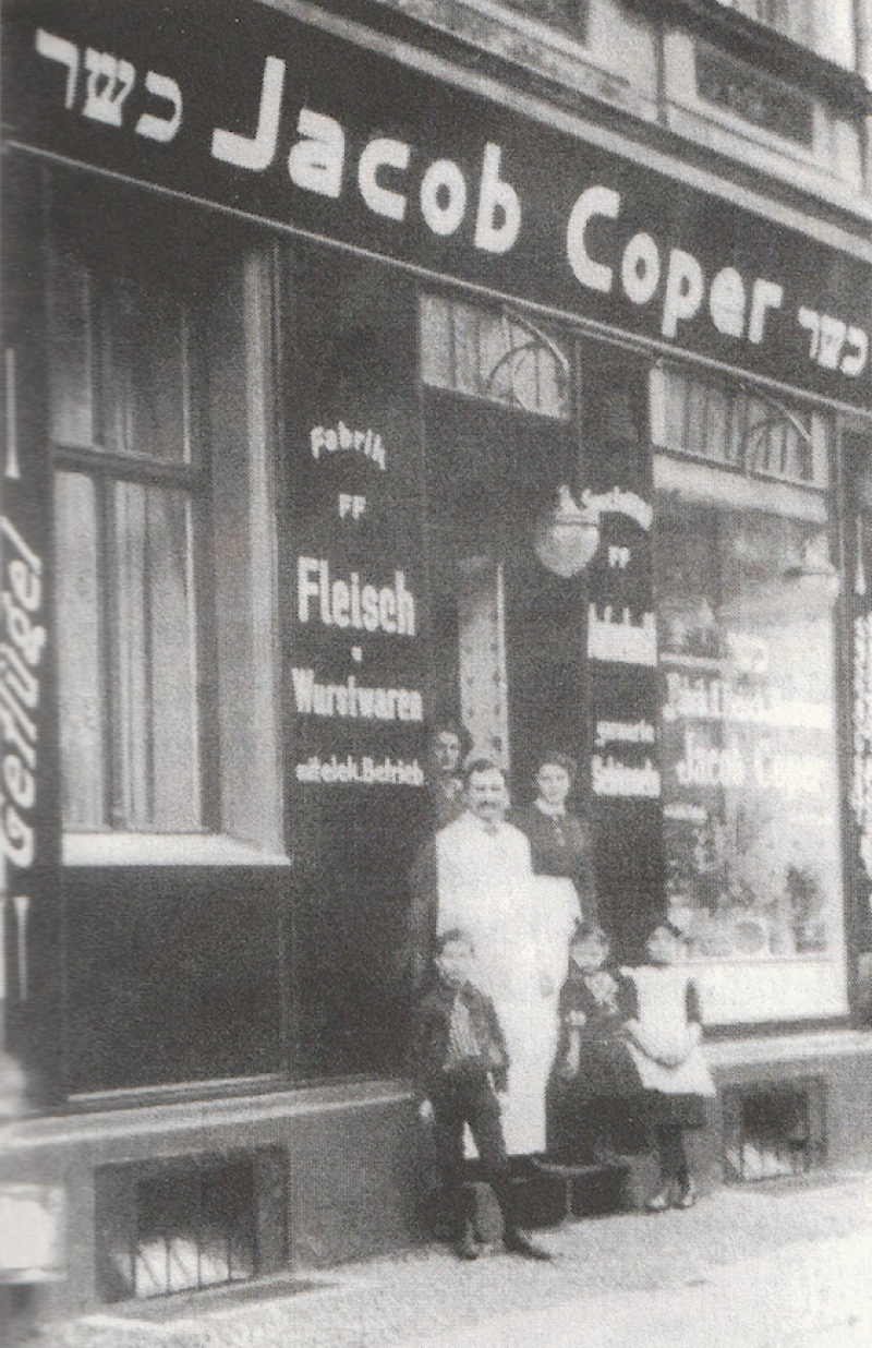 Eine schwarz-weiß Aufnahme des Geschäfts von Jacob Coper, ca. 1920. Drei Kinder, der Geschäftsinhaber und zwei Frauen stehen am Eingan des Geschäfts. Auf den Geschäftsladen sind Inschriften zu lesen: Oberhalb der Tür "Jacob Coper" und neben dem Eingang "Fabrik FF Fleich und Wurtstwaren mit elekt. Betrieb"