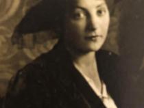 Die monochrome Fotografie in Sepiatönen zeigt Anna le Coutre. Ein junge Frau mit Hut und welligem Harr, die auf einem Stuhl sitzend in die Kamera schaut. Die Fotografie stammt etwa aus dem Jahr 1916.