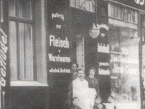 Eine schwarz-weiß Aufnahme des Geschäfts von Jacob Coper, ca. 1920. Drei Kinder, der Geschäftsinhaber und zwei Frauen stehen am Eingan des Geschäfts. Auf den Geschäftsladen sind Inschriften zu lesen: Oberhalb der Tür &amp;quot;Jacob Coper&amp;quot; und neben dem Eingang &amp;quot;Fabrik FF Fleich und Wurtstwaren mit elekt. Betrieb&amp;quot;