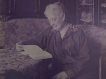 Elise Gutkind sitzt an einem Tisch in einem gutbürgerlichen Wohnzimmer und liest in einem Buch